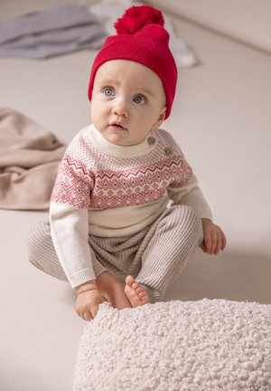 Gestrickter Pullover mit rotem geometrischem Muster, cremefarbener Basis und Knopfakzenten, kombiniert mit grauen gerippten Hosen; rote Bommelmütze; strukturierter Kissen.