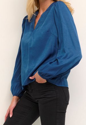 Blaue Bluse mit V-Ausschnitt, langen Puffärmeln und lockerer Passform. Der Stoff hat eine glatte, leicht glänzende Textur. Kombiniert mit schwarzen Hosen.