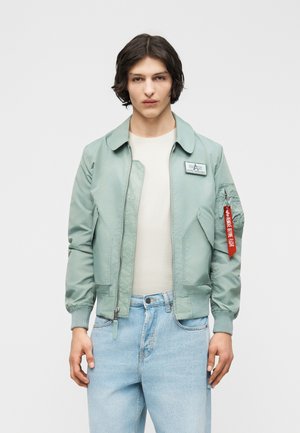 Junger Mann trägt eine hellgrüne Bomberjacke mit Reißverschluss, rotem Etikett am Ärmel, weißem Hemd und hellblauen Jeans vor einfarbigem Hintergrund.