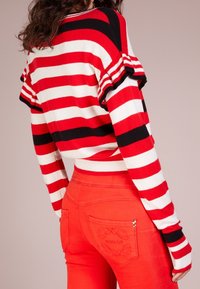 Pull en maille rayé rouge et blanc avec manches courtes à volants, associé à un jean rouge ajusté avec poches arrière texturées et détails de couture.