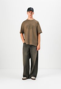 En ung mann med svart caps, en overdimensjonert brun t-skjorte med "Dry Goods" trykk, vide falmede svarte jeans og hvite sneakers, står foran en hvit bakgrunn.