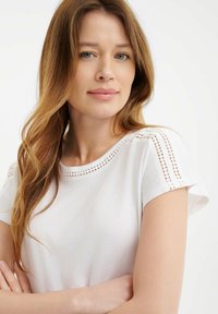 Biała koszulka typu t-shirt z krótkim rękawem i okrągłym dekoltem, z wplecionymi koronkowymi detalami na ramionach, wykonana z miękkiej tkaniny o gładkiej fakturze.