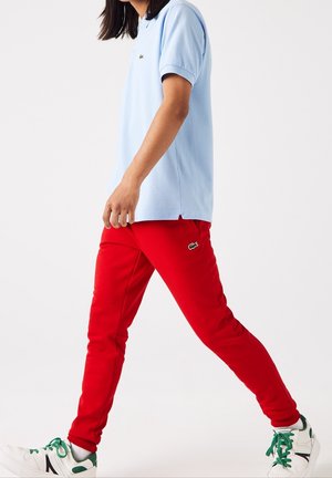 Eine Person, die ein hellblaues Poloshirt, rote Hosen mit Logo und weiße Turnschuhe mit grünen Schnürsenkeln vor einem weißen Hintergrund trägt.
