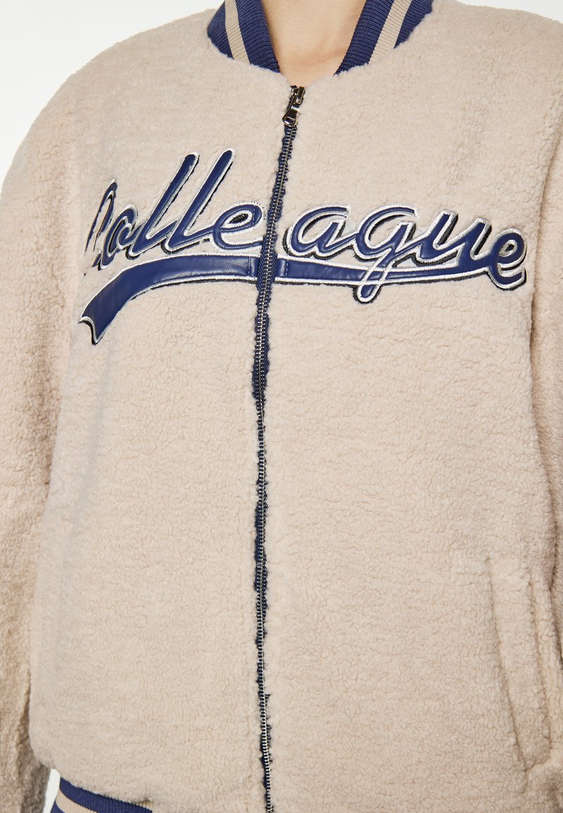 Beige Bomberjacke mit Sherpa-Futter und marineblauen Akzenten. Verfügt über einen gerippten Kragen, einen Frontreißverschluss und gestickten "Kollege"-Text. Zwei Seitentaschen.