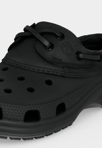 Μαύρο λαστιχένιο τσόκαρο με οπές αερισμού, υφασμένη σόλα και ρυθμιζόμενο κορδόνι σε απλό ανοιχτό γκρι φόντο.