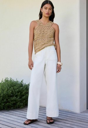 Femme debout sur une terrasse en bois portant un haut sans manches beige en crochet, un pantalon blanc à jambes larges, des sandales marron et un bracelet jonc.