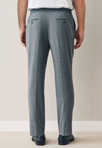 Graue, maßgeschneiderte Hose aus strukturiertem Stoff, mit zwei aufgesetzten Gesäßtaschen mit Knöpfen und flachem Vorderteil. Kombiniert mit schwarzen Schuhen.