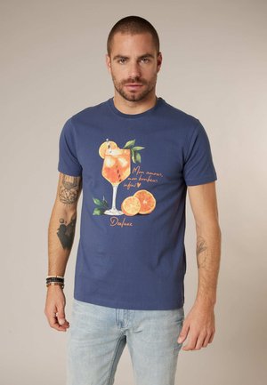 Homme aux cheveux courts et tatouages, portant un t-shirt bleu avec un motif de cocktail orange et des tranches d'orange, et un jean clair, debout.