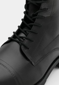 Botín de cuero negro hasta el tobillo con una textura suave, que presenta ojales y cordones tradicionales. Punta estructurada y costuras detalladas a lo largo de las uniones.