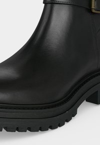 Primer plano de una bota de tobillo de cuero negro con tacón grueso y suela con estrías gruesas para mayor agarre y durabilidad.