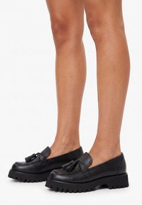Mocasines de cuero negro con borlas y suelas gruesas de taco usados sobre piernas desnudas contra un fondo blanco.
