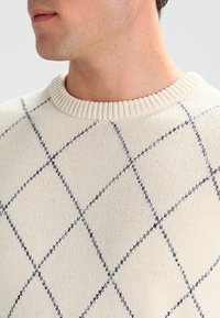 Maglione a maglia beige con collo a giro ribs, caratterizzato da un motivo a diamante blu navy sulla parte anteriore, creando un design geometrico e testurizzato.