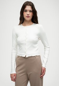 Biały prążkowany kardigan z długim rękawem, okrągłym dekoltem, zapięciem na guziki z przodu oraz falbaniastym wykończeniem u dołu i na mankietach. Połączony z beżowymi spodniami.