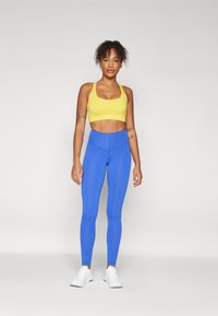 Sárga sportmelltartó vastag pántokkal és négyzet alakú nyakkivágással, magas derékú kék leggings-szel párosítva. A modellen fehér sportcipők vannak.
