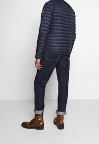 Mørkeblå vatteret jakke med vandret syning, kombineret med mørke denimbukser og brune snørestøvler. Oprullede manchetter på jeans.