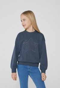 Tmavě modrá mikina s velkým, texturovaným hvězdným designem na přední straně. Má pružné manžety a lem, s uvolněným střihem. V kombinaci s modrými džíny.