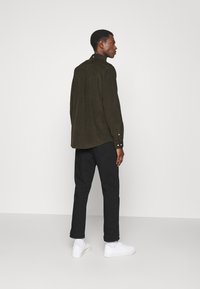 Donkergroen suède shirt met een kraag, knoopmanchetten en een ronde zoom, gecombineerd met zwarte broeken en witte sneakers. Minimalistisch design.