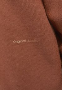 Textil marrón con una textura suave y de punto, con un sutil brillo; incluye la marca en relieve "Originals Studio".