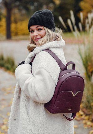 Bordeauxfarbener Rucksack aus strukturiertem Material mit hexagonalem Muster; verfügt über einen V-förmigen Akzent und verstellbare Träger. Getragen über einem weißen Mantel.