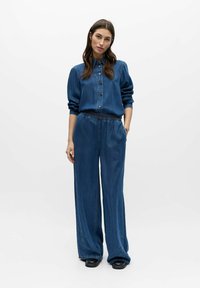 Femme debout portant une chemise en denim bleue à boutons et un pantalon à jambes larges avec broderies florales près des chevilles, mains dans les poches, fond neutre.