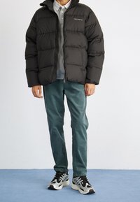 Sort pufferjakke med lynlås og hætte, parret med en lys grå sweatshirt, teal farvede corduroy bukser og sorte sneakers med hvide accenter.