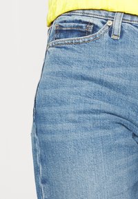 Jeans de ganga azul claro com um corte justo, apresentando um design clássico de cinco bolsos e costura bege. O tecido tem uma aparência macia e ligeiramente texturizada.