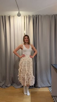 Nainen seisoo käsivarret lanteilla, yllään valkoinen hihaton yläosa ja hulmuava beige kukkakuosinen hame huoneessa, jossa on harmaat verhot ja puulattia.