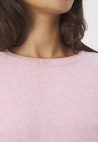 Ljusrosa stickad tröja med ribbad rund halsringning, mjuk textur och raglanärmar. Tyget ser slätt ut med subtila variationer.