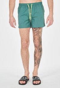 Costumi da bagno verdi con finitura testurizzata, vita elastica e cordoncino giallo. Infradito nere con logo bianco sulla parte anteriore.