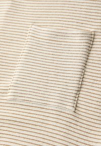 Nahaufnahme eines beigen und weiß gestreiften Stoffs mit einer kleinen, rechteckigen Tasche, die aufgenäht ist und zum Muster des Stoffs passt.