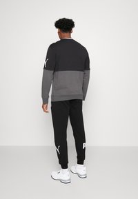 Černá a šedá sportovní bunda s dlouhými rukávy, spárovaná s černými tepláky. Obsahuje loga Puma a má volný střih. Bílá sneakers dotvářejí celý vzhled.