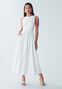 Balta maxidress su tekstūruotu liemeniu, be rankovių dizainu ir platėjančia sijoneliu. Turi šonines kišenes ir derinama su austi beige aukštakulniais.