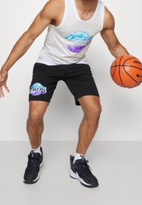 Czarne spodenki sportowe z kolorową grafiką "Los Angeles Lakers", połączone z białym topem na ramiączkach i czarnymi butami sportowymi, kozłując piłkę koszykową.