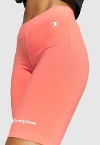 Detailní záběr osoby v korálově růžových cyklonohavicích Champion s logem na stehně a na pásu na pozadí bez vzoru.