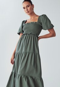 Zelena haljina s višestrukim slojevima, napuhanim rukavima, i naboranom bustierom, ukrašena naborima. Napravljena od mekog materijala s glatkom teksturom i minimalnim uzorkom.