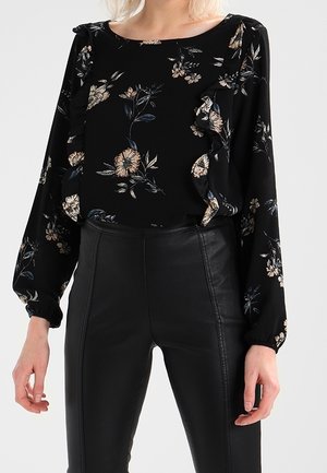 Schwarze Langarmbluse mit beigem Blumenmuster und Rüschen-Details, kombiniert mit schwarzen Lederhosen mit hoher Taille.