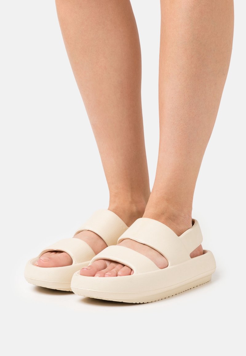 Sandales couleur crème avec deux larges brides sur le pied, design ouvert à l'avant, semelle épaisse et finition texturée pour plus d'adhérence.