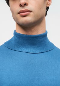 Μπλε πουλόβερ με λαιμόκοψη ζιβάγκο, κατασκευασμένο από απαλό, πλεκτό ύφασμα, με ψηλό λαιμό και επίπεδη γιακά. Η υφή είναι λεία και το σχέδιο εφαρμοστό.