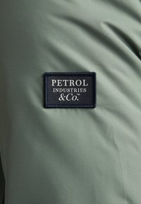 Hellgrüner Stoff mit strukturierter Oberfläche zeigt ein rechteckiges, marineblaues Etikett mit der weißen Aufschrift "PETROL INDUSTRIES & CO.".