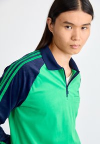 Zeleni i tamnoplavi pulover s patentnim zatvaračem, dugim rukavima, s kontrastnim prugama i džepom na prsima. Materijal izgleda gladak i lagan.