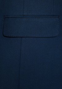 Granatowy materiał o teksturowanej powierzchni. Zawiera kwadratową kieszeń naszywaną bez widocznych elementów metalowych czy ozdób. Czyste, proste linie.