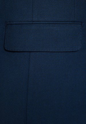 Granatowy materiał o teksturowanej powierzchni. Zawiera kwadratową kieszeń naszywaną bez widocznych elementów metalowych czy ozdób. Czyste, proste linie.