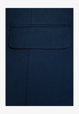 Granatowy materiał o teksturowanej powierzchni. Zawiera kwadratową kieszeń naszywaną bez widocznych elementów metalowych czy ozdób. Czyste, proste linie.