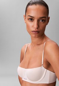 Lichtbeige bralette met dunne bandjes, naadloze cups en een klein merkembleem aan de voorkant, met een gladde textuur en eenvoudig ontwerp.