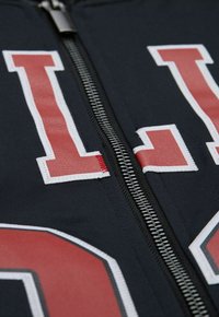 Marine-blaues Zip-Jackett mit großen weißen und roten Zahlen, sichtbarem Metallreißverschluss und strukturiertem Stoff; Schnittdetails an der Schrift.