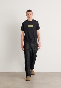 Svart T-skjorte med rund hals og gult-grønt "calvin klein"-logo, kombinert med mørk grå vide bukser og lys brune sko.
