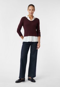 Frau steht und trägt einen burgunderfarbenen Strickpullover über einem weißen Hemd mit Kragen, dunkle weit geschnittene Jeans und schwarze Loafers vor einem schlichten Hintergrund.