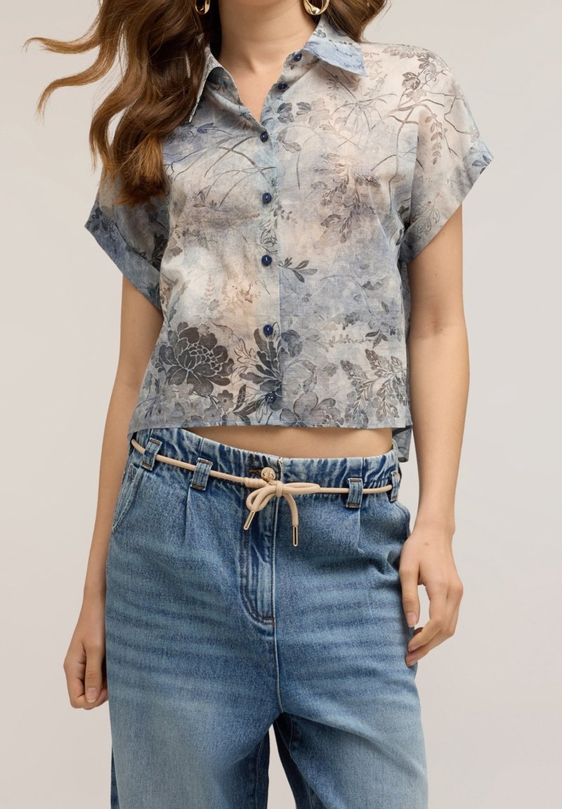 Femme portant une chemise fleurie à manches courtes boutonnée, portée sortie sur un jean bleu taille haute avec une ceinture en corde beige nouée devant.