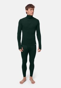 Uomo che indossa un intimo termico aderente verde scuro con collo a zip corta e pantaloni aderenti, in piedi a piedi nudi su sfondo bianco.