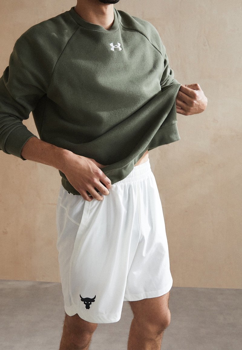 Homme portant un sweat Under Armour vert olive, remontant l'ourlet, short blanc avec logo de taureau noir, fond beige.
